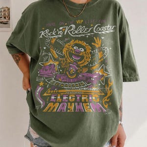Puede incluir: Camiseta verde oliva con un dise&ntilde;o gr&aacute;fico de un personaje de los Muppets y el texto "Rock n' Roller Coaster Featuring Electric Mayhem". El dise&ntilde;o incluye detalles en morado, amarillo y blanco. La camiseta tiene mangas cortas y un corte holgado.