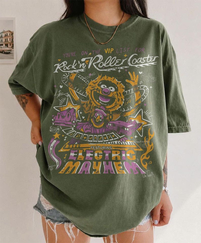 Puede incluir: Camiseta verde oliva con un dise&ntilde;o gr&aacute;fico de un personaje de los Muppets y el texto "Rock n' Roller Coaster Featuring Electric Mayhem". El dise&ntilde;o incluye detalles en morado, amarillo y blanco. La camiseta tiene mangas cortas y un corte holgado.