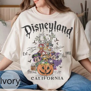 May include: Ivory t-shirt with a Disneyland graphic featuring Halloween-themed Disney characters such as Mickey and Minnie Mouse. The design includes the text "Disneyland California" and the year "1955".