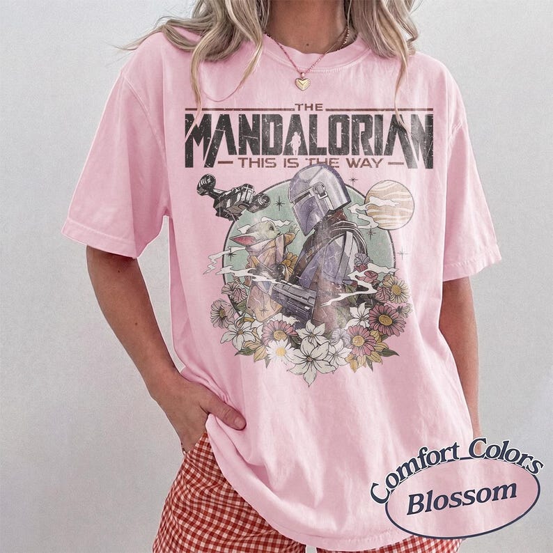 May include: A light pink short-sleeve t-shirt featuring a graphic print of The Mandalorian and Grogu. The text reads THE MANDALORIAN THIS IS THE WAY. The design includes a spaceship, a planet, and various flowers in a circular frame on the chest.