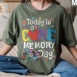 以下が含まれることがあります： 緑色のTシャツに、カラフルなグラフィックがプリントされています。「Today is a CORE MEMORY Day」という文字と、COREの文字の中にミッキーマウスの頭が描かれています。グラフィックには、映画「インサイド・ヘッド」のキャラクターも描かれています。