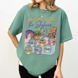 May include: Green t-shirt with a graphic featuring the text "WELCOME ABOARD The Skyliner" and images of Disney characters in gondolas. The design includes illustrations of Epcot's Spaceship Earth, Cinderella's Castle, and the Hollywood Tower Hotel.