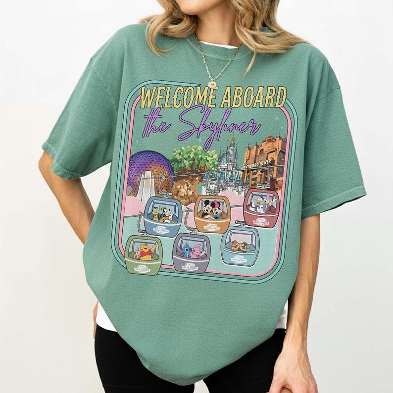 May include: Green t-shirt with a graphic featuring the text "WELCOME ABOARD The Skyliner" and images of Disney characters in gondolas. The design includes illustrations of Epcot's Spaceship Earth, Cinderella's Castle, and the Hollywood Tower Hotel.