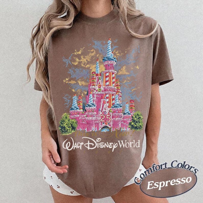 May include: Espresso-colored t-shirt featuring a pink castle graphic with blue and yellow accents, the number 25, and the words "Walt Disney World." The shirt is part of the Comfort Colors collection.