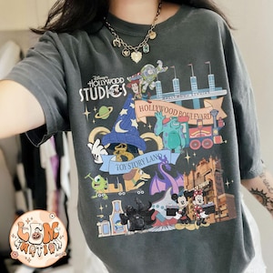 Può includere: T-shirt grigio scuro con una grafica colorata di Disney's Hollywood Studios, con personaggi di Toy Story, Monsters, Inc. e Topolino e Minni. La maglietta presenta il testo "Hollywood Studios" e "Toy Story Land".