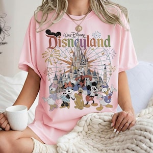 May include: Pink t-shirt featuring a Disneyland graphic. The design includes the Disneyland logo, a castle, fireworks, and cartoon characters such as Mickey Mouse, Minnie Mouse, Donald Duck, and Goofy. The shirt is short-sleeved.