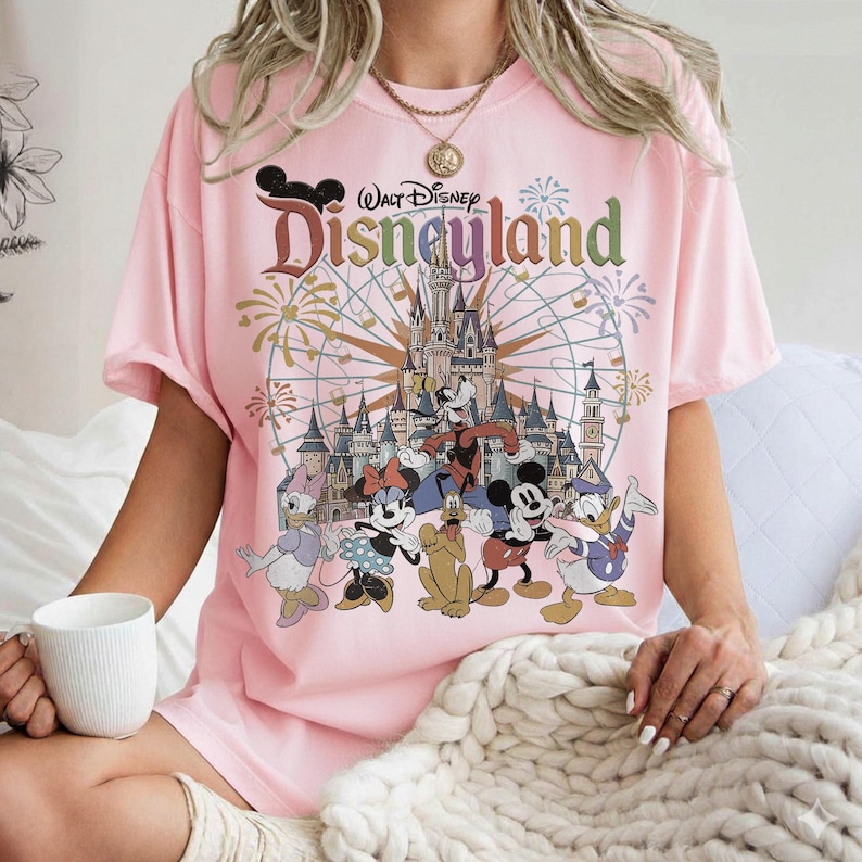 May include: Pink t-shirt featuring a Disneyland graphic. The design includes the Disneyland logo, a castle, fireworks, and cartoon characters such as Mickey Mouse, Minnie Mouse, Donald Duck, and Goofy. The shirt is short-sleeved.