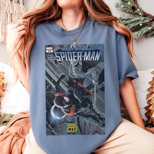 Op de afbeelding: Een lichtblauw T-shirt met een Miles Morales Spider-Man stripboekcover. De cover toont Spider-Man in een dynamische pose met een zwart en rood pak, met de woorden "GANG WAR IS COMING".