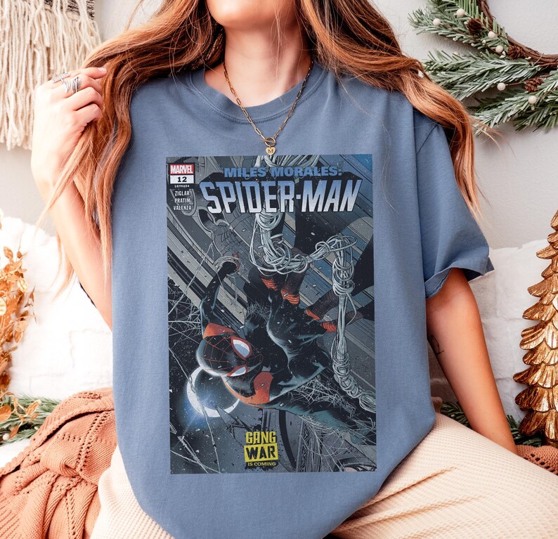 Op de afbeelding: Een lichtblauw T-shirt met een Miles Morales Spider-Man stripboekcover. De cover toont Spider-Man in een dynamische pose met een zwart en rood pak, met de woorden "GANG WAR IS COMING".