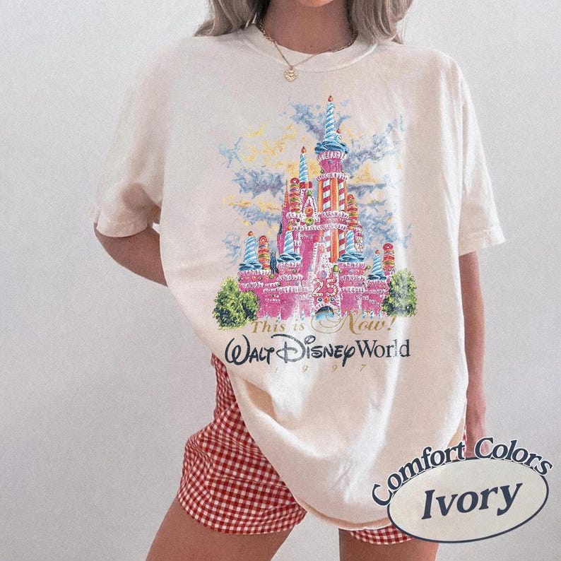 May include: Ivory-colored t-shirt featuring a colorful graphic of a pink castle with blue turrets, green bushes, and the text "This is Now! Walt Disney World 1997." The shirt is from Comfort Colors.