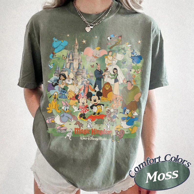 May include: A moss green short-sleeve t-shirt featuring a large, colorful graphic print of various animated characters and a castle. The text "Magic Kingdom" and "Walt Disney World" is printed in red and white at the bottom of the graphic design.