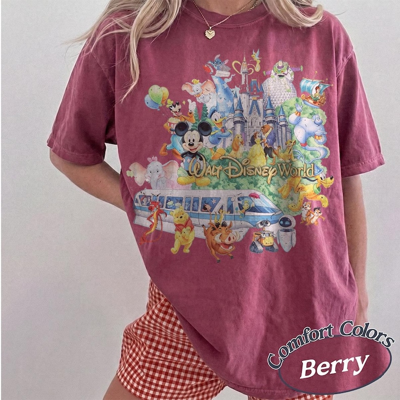 May include: A berry-colored short-sleeve t-shirt featuring a large graphic print of Walt Disney World with various animated characters, a monorail, and the castle. The shirt has a crew neckline and a relaxed fit, showcasing a vintage-style wash.