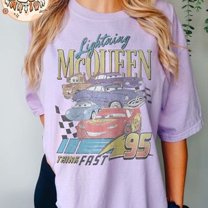 May include: A lavender t-shirt with a vintage-style graphic of Lightning McQueen from the Cars movie. The graphic features the text "Lightning McQueen" and "Think Fast" along with the number "95".