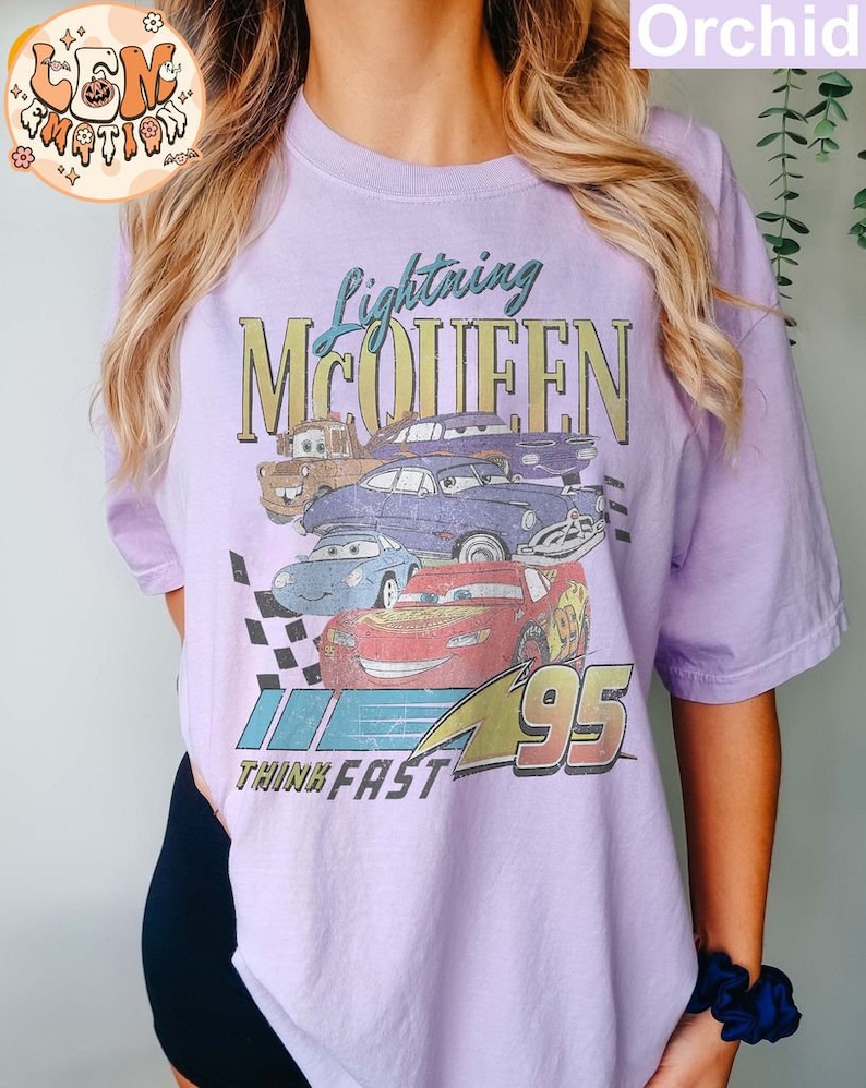 May include: A lavender t-shirt with a vintage-style graphic of Lightning McQueen from the Cars movie. The graphic features the text "Lightning McQueen" and "Think Fast" along with the number "95".