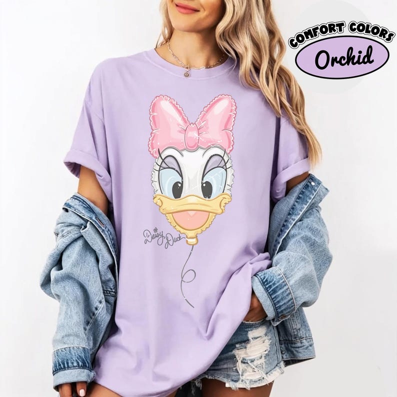 Pode incluir: Uma t-shirt cor de orqu&iacute;dea com um gr&aacute;fico da Daisy Duck desenhado como um bal&atilde;o cor-de-rosa com um la&ccedil;o grande. A t-shirt inclui o texto Daisy Duck em letra cursiva abaixo do gr&aacute;fico do bal&atilde;o e uma etiqueta Comfort Colors Orchid.