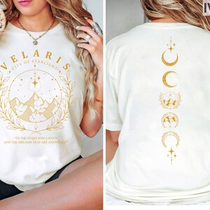 May include: Ivory-colored t-shirt with gold text and graphics. The front features the words "VELARIS CITY OF STARLIGHT" with a mountain and star design. The back has a vertical design of moons, stars, and mountain graphics. The text "TO THE STARS WHO LISTEN AND THE DREAMS THAT ARE ANSWERED" is also visible.