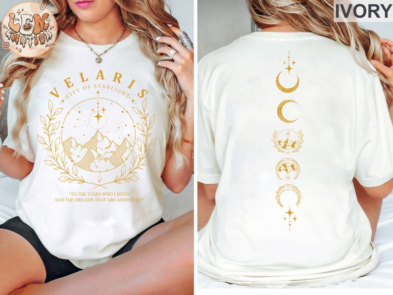 May include: Ivory-colored t-shirt with gold text and graphics. The front features the words "VELARIS CITY OF STARLIGHT" with a mountain and star design. The back has a vertical design of moons, stars, and mountain graphics. The text "TO THE STARS WHO LISTEN AND THE DREAMS THAT ARE ANSWERED" is also visible.