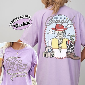 May include: A light purple t-shirt featuring a back graphic of a cowgirl with a braided ponytail, a red hat, a yellow vest, and a cow-print boot with a snake. The front graphic displays the text "It's a Toy Story baby just yes say" in rope font.