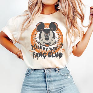 May include: A white t-shirt with a vintage-style graphic of Mickey Mouse with fangs. The graphic is in a circle with the text "Mickey Mouse Fang Club" around it.