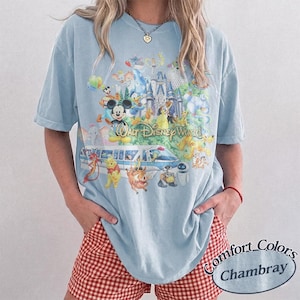 May include: A light blue short-sleeve t-shirt featuring a colorful graphic print of Walt Disney World with various animated characters, a monorail, and a castle. The shirt has a crew neckline and a relaxed fit, showcasing a detailed collage design.