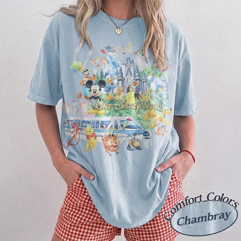 May include: A light blue short-sleeve t-shirt featuring a colorful graphic print of Walt Disney World with various animated characters, a monorail, and a castle. The shirt has a crew neckline and a relaxed fit, showcasing a detailed collage design.