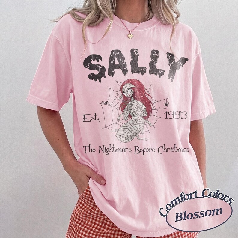 May include: A light pink short-sleeve t-shirt featuring a graphic of the character Sally from The Nightmare Before Christmas. The design includes a spiderweb, the text SALLY, Est. 1993, and The Nightmare Before Christmas in black ink.