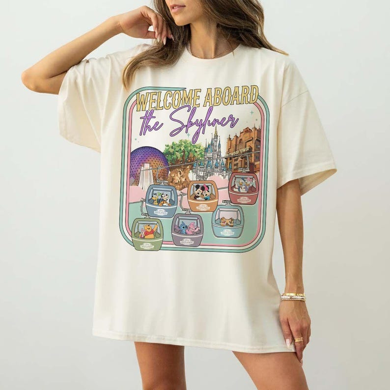 May include: Cream-colored t-shirt featuring a colorful graphic with the text "WELCOME ABOARD The Skyliner." The graphic includes cartoon characters in gondolas, with a theme park backdrop. The shirt has short sleeves and a relaxed fit.