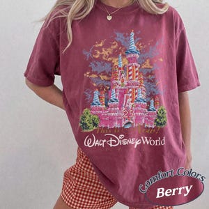 May include: Berry-colored t-shirt featuring a graphic of a pink castle with blue and orange accents, and the text "Walt Disney World" and "This is Now!" The shirt also has the text "Comfort Colors Berry".