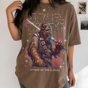 K&ouml;nnte beinhalten: Braunes T-Shirt mit einer Grafik von Chewbacca, der ein blaues Lichtschwert h&auml;lt, dem Star Wars-Logo und dem Text "ATTACK OF THE CLONES". Das Shirt hat einen Vintage-Look.