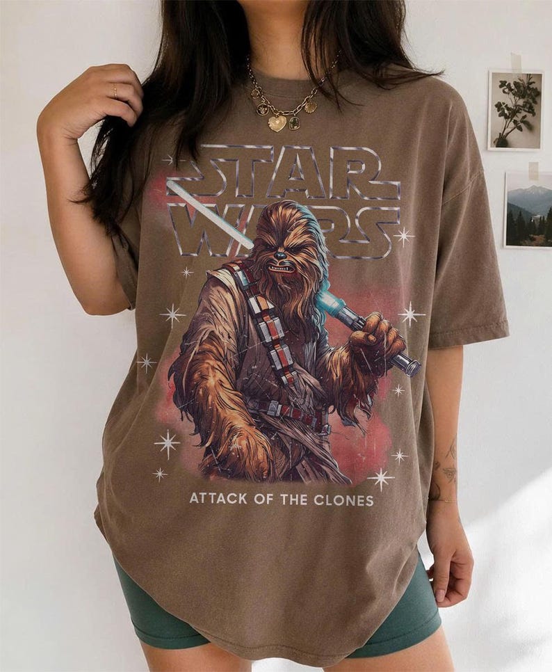 K&ouml;nnte beinhalten: Braunes T-Shirt mit einer Grafik von Chewbacca, der ein blaues Lichtschwert h&auml;lt, dem Star Wars-Logo und dem Text "ATTACK OF THE CLONES". Das Shirt hat einen Vintage-Look.