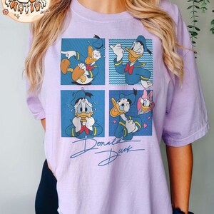 May include: A purple t-shirt with a cartoon image of Donald Duck in four different expressions. The shirt has the text "Donald Duck" written in a stylized font.