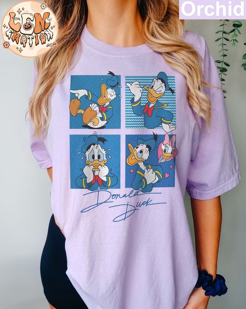 May include: A purple t-shirt with a cartoon image of Donald Duck in four different expressions. The shirt has the text "Donald Duck" written in a stylized font.
