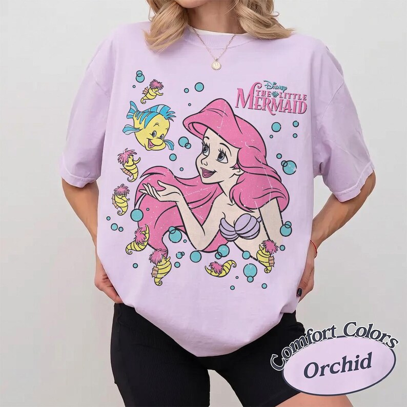 May include: A light purple short-sleeve t-shirt featuring a graphic print of Ariel from The Little Mermaid with pink hair, a purple seashell top, and Flounder. The design includes small yellow sea creatures and blue bubbles with the text Disney The Little Mermaid.