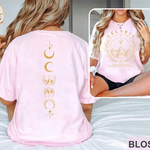 May include: Light pink t-shirt with gold graphic designs. The back features a vertical design of moons and stars. The front has the text "VELARIS CITY OF STARLIGHT" with a mountain and star design. The word "BLOSSOM" is at the bottom.