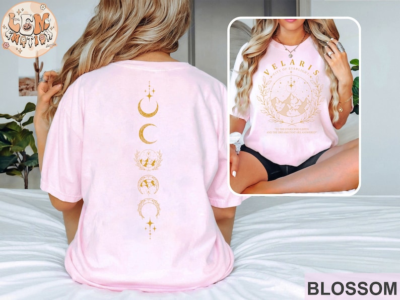 May include: Light pink t-shirt with gold graphic designs. The back features a vertical design of moons and stars. The front has the text "VELARIS CITY OF STARLIGHT" with a mountain and star design. The word "BLOSSOM" is at the bottom.