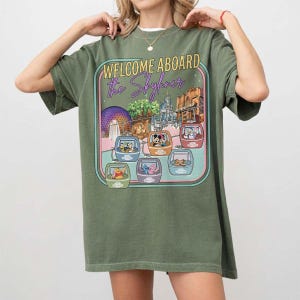 May include: Olive green t-shirt featuring a graphic with the text "WELCOME ABOARD the Skyliner." The graphic shows various Disney characters in gondolas, with park landmarks in the background. The shirt is a relaxed fit.