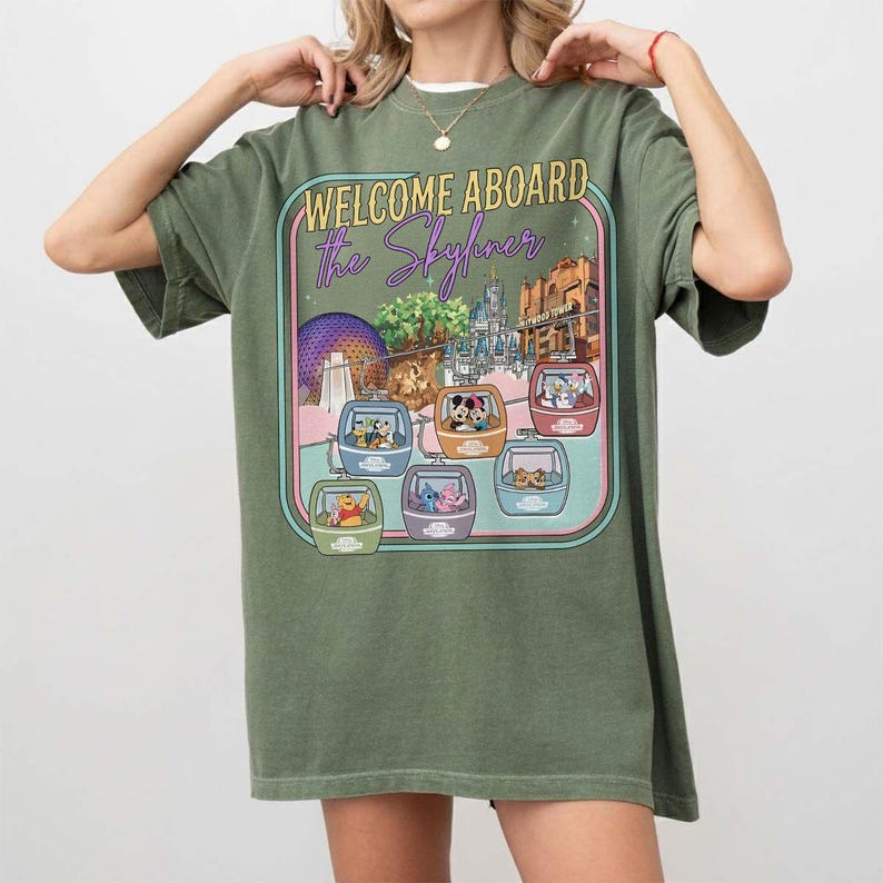 May include: Olive green t-shirt featuring a graphic with the text "WELCOME ABOARD the Skyliner." The graphic shows various Disney characters in gondolas, with park landmarks in the background. The shirt is a relaxed fit.