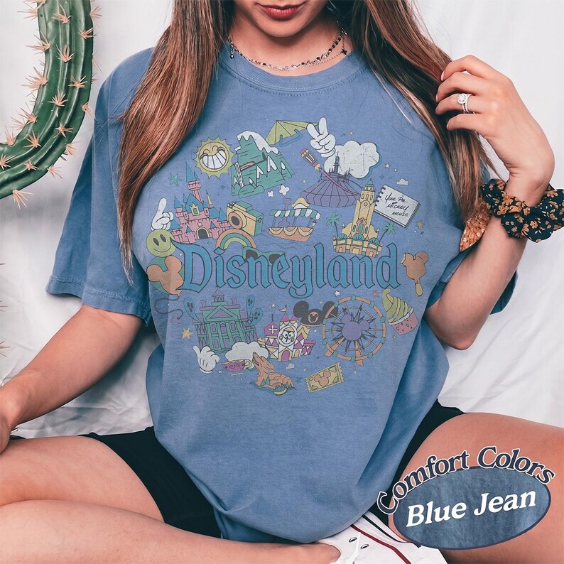 May include: Blue jean colored t-shirt with a Disneyland graphic. The graphic includes various park icons, such as a castle, a Ferris wheel, and Mickey Mouse. The words "Disneyland" are written in a whimsical font.