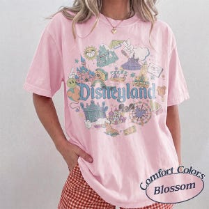 May include: A light pink t-shirt with a Disneyland graphic. The graphic features various park icons and the word "Disneyland" in a whimsical font. The shirt is labeled "Comfort Colors Blossom".
