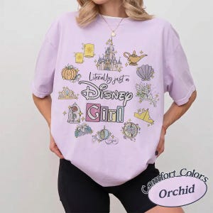 Op de afbeelding: Een lichtpaars T-shirt met de tekst "Literally just a Disney Girl" omringd door Disney-thema graphics. Het shirt heeft ook de woorden "Comfort Colors" en "Orchid" in een cirkelvormig ontwerp.