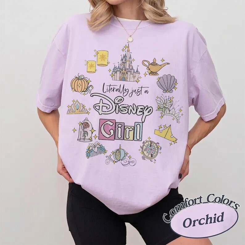 Op de afbeelding: Een lichtpaars T-shirt met de tekst "Literally just a Disney Girl" omringd door Disney-thema graphics. Het shirt heeft ook de woorden "Comfort Colors" en "Orchid" in een cirkelvormig ontwerp.