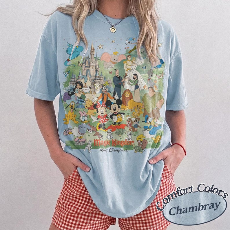 May include: A light blue short-sleeve t-shirt featuring a large, colorful graphic print of various animated characters, a castle, and the text Magic Kingdom Walt Disney World. The shirt is made by Comfort Colors in the color Chambray.