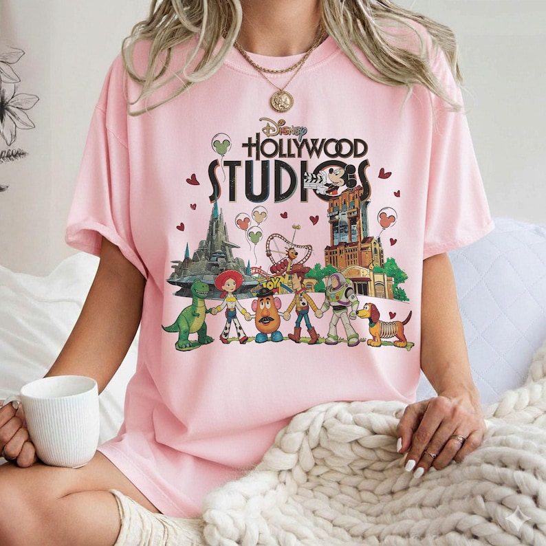Op de afbeelding: Een lichtroze T-shirt met het Disney Hollywood Studios-logo en tekenfilmfiguren. De afbeelding bevat Mickey Mouse, een dinosaurus, Woody, Buzz Lightyear en andere personages. Het shirt heeft korte mouwen.