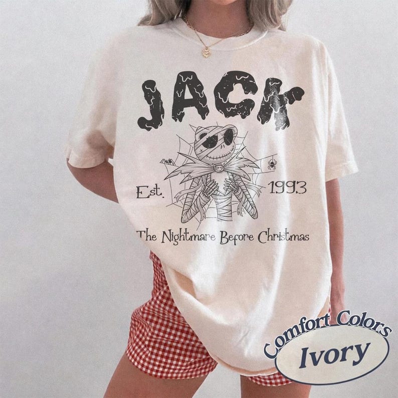 May include: An ivory short-sleeve t-shirt featuring a black graphic of a skeletal character with stitched details, a spiderweb background, and the text "JACK", "Est. 1993", and "The Nightmare Before Christmas" printed in a distressed black font.