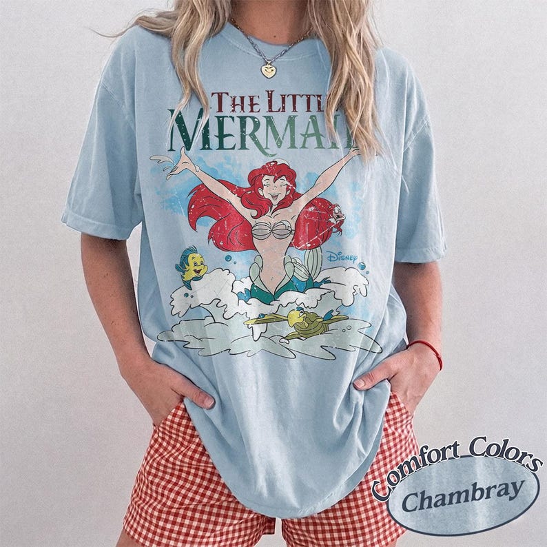 May include: A light blue short-sleeve t-shirt featuring a graphic print of Ariel from The Little Mermaid with red hair, a seashell top, and a green tail, surrounded by Flounder and Sebastian, with the text The Little Mermaid and Disney.