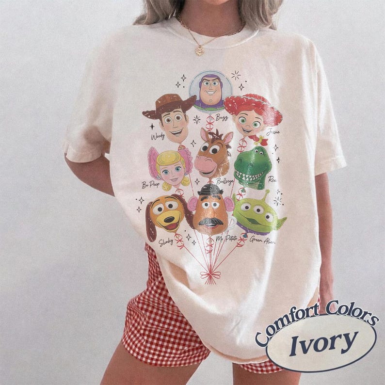 以下が含まれることがあります： アイボリーの半袖Tシャツで、風船として描かれた様々なアニメキャラクターのグラフィックプリントが特徴です。キャラクターには、ウッディ、バズ、ジェシー、ボー・ピープ、ブルズアイ、レックス、スリンキー、ミスター・ポテトヘッド、緑色のエイリアンが含まれており、赤い紐で繋がっています。