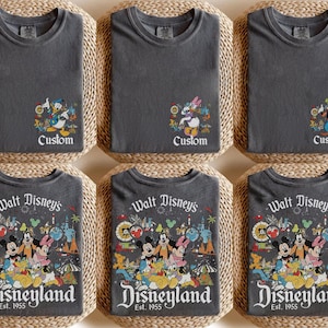 Puede incluir: Seis camisetas grises con dise&ntilde;os de personajes de Disney. Tres camisetas tienen el texto "Custom" con el Pato Donald, Daisy y Goofy. Las otras tres camisetas dicen "Walt Disney's Disneyland Est. 1955" con Mickey, Minnie y otros personajes.