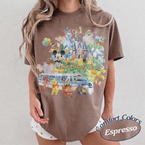 May include: A brown short-sleeve t-shirt featuring a graphic print of various animated characters, a castle, and a monorail train. The text "Walt Disney World" is printed in gold script across the center of the design. The shirt has a crew neckline.
