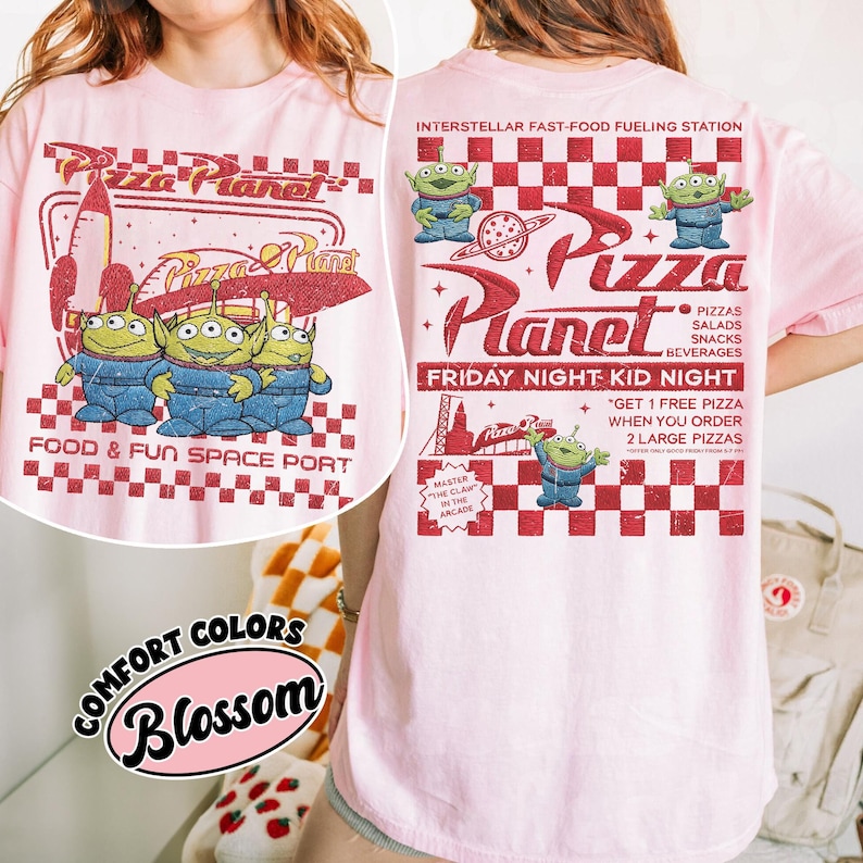 K&ouml;nnte beinhalten: Hellrosa T-Shirt mit Pizza Planet-Design. Auf der Vorderseite sind drei gr&uuml;ne Aliens in blauen Overalls, eine Rakete und der Text "Food & Fun Space Port" abgebildet. Auf der R&uuml;ckseite befinden sich das Pizza Planet-Logo, Aliens und Text.