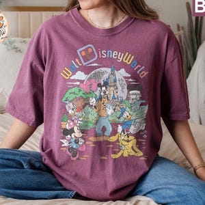 May include: A purple t-shirt with a vintage-style graphic of Mickey Mouse and friends at Walt Disney World. The graphic features the text "Walt Disney World" in a colorful, retro font.